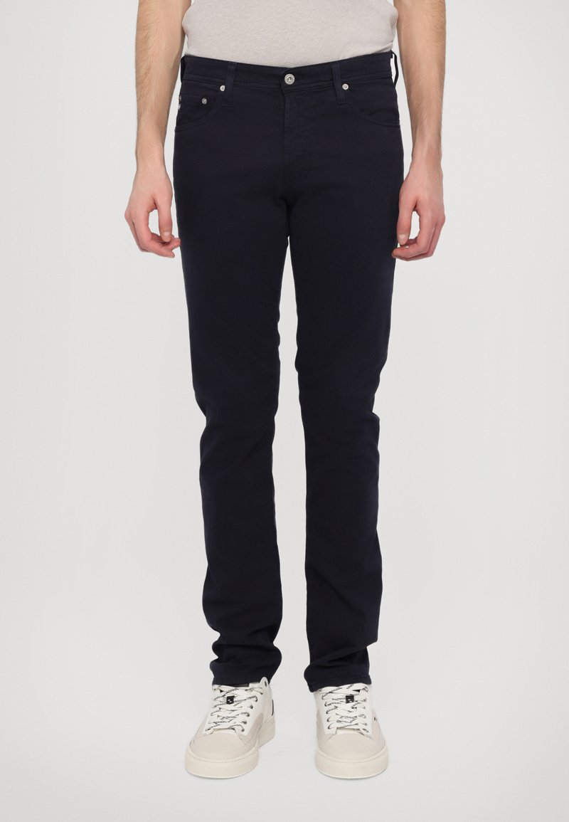 Σκούρο μπλε υφασμάτινα τζιν σε λεπτή γραμμή με πέντε τσέπες, μπροστινό κλείσιμο με κουμπί και ελάχιστες λεπτομέρειες ραφής. Φορεμένα με λευκά αθλητικά παπούτσια.