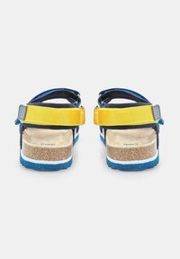 Sandalite tagumine vaade, millel on tumesinised rihmad, kollane reguleeritav rihm, korkjalats ja sinine kummitald valgete aktsentidega.