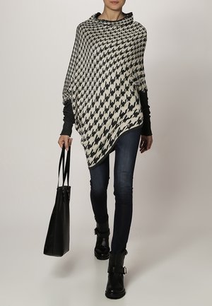 Poncho a scacchi bianco e nero con collo a cowl, abbinato a jeans skinny blu scuro e stivaletti neri alla caviglia, mentre porta una borsa a tracolla nera.