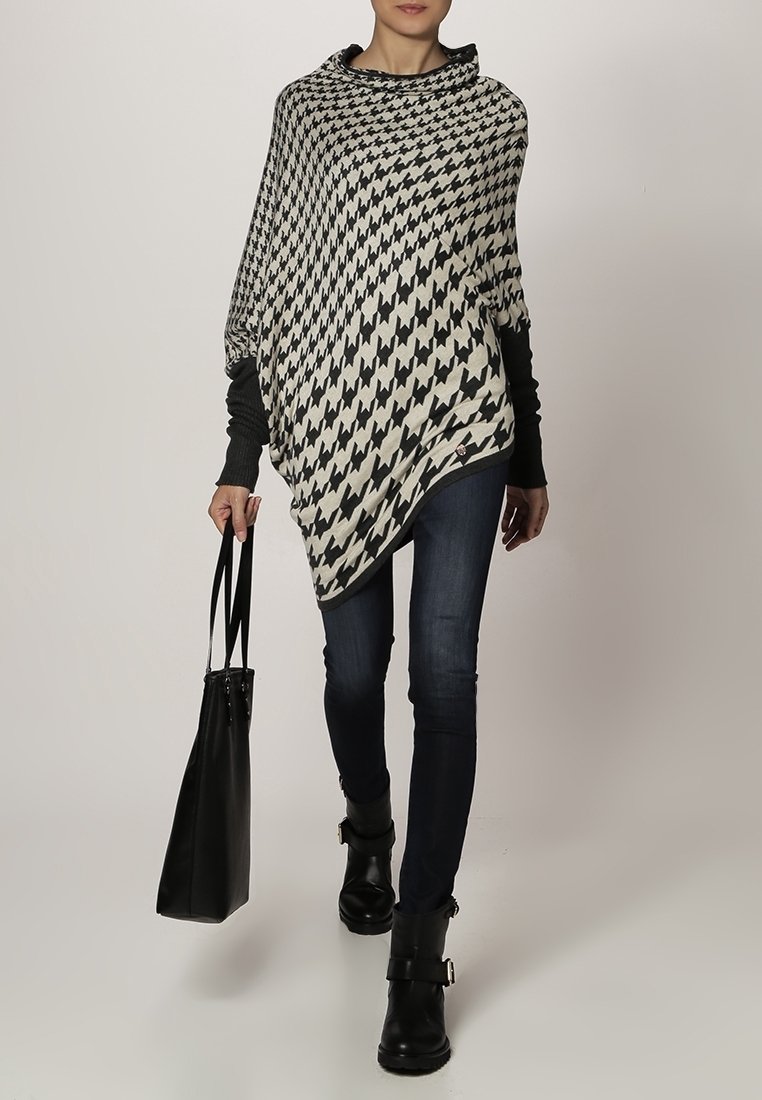 Zwart-wit houndstooth-patroon poncho met een coltraject, gecombineerd met donkerblauwe skinny jeans en zwarte enkellaarsjes, terwijl ze een zwarte tote bag draagt.