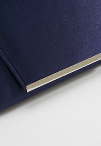 Pochette en tissu bleu marine avec une finition métallique argentée élégante sur le rabat, sur un fond gris clair.