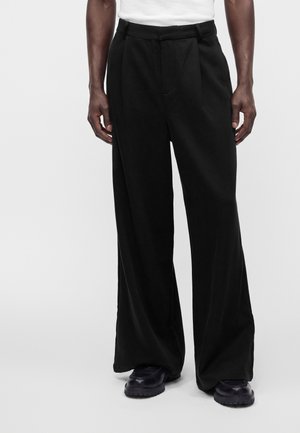 Personne portant un pantalon noir à jambes larges et des chaussures noires, debout les mains détendues le long du corps sur un fond clair uni.