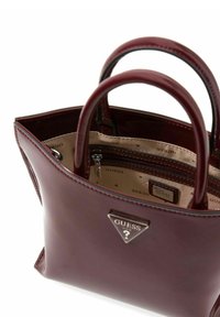 Bordeauxleren handtas met dubbele handvatten, driehoekig zilveren logo en beige interieur met "GUESS"-patroon en een vak met rits.