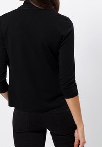 Schwarzes, eng anliegendes Shirt mit dreiviertel Ärmeln, glatter Textur und hohem Kragen. Nahtdetails auf der Rückseite. Getragen mit schwarzer Hose.