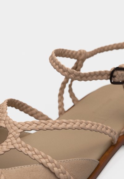 Μπεζ φλατ σανδάλι με πλεγμένους ιμάντες και μαύρη πόρπη σε ουδέτερο φόντο.