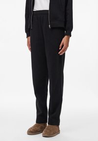 Juodi treningai su elastingu juosmeniu, siaurėjančiomis kojomis ir šoninėmis kišenėmis. Kombinuojami su tan spalvos zomšos šlepetėmis.