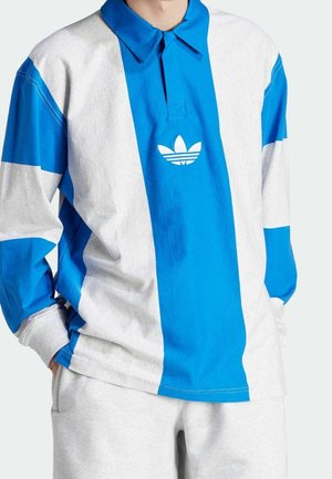 Homme portant un maillot de rugby à manches longues avec de larges rayures verticales blanches et bleues et un logo tréflé Adidas blanc centré sur la poitrine.