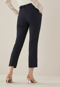 Donna che indossa pantaloni navy a vita alta con tasche posteriori e mule beige con tacco, in piedi di fronte a uno sfondo chiaro e semplice.