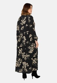Dlouhá černá šaty s béžovým květinovým vzorem, dlouhé rukávy, plisované detaily a splývavá silueta. Nosené s černými kozačkami na vysokém podpatku až po kolena.