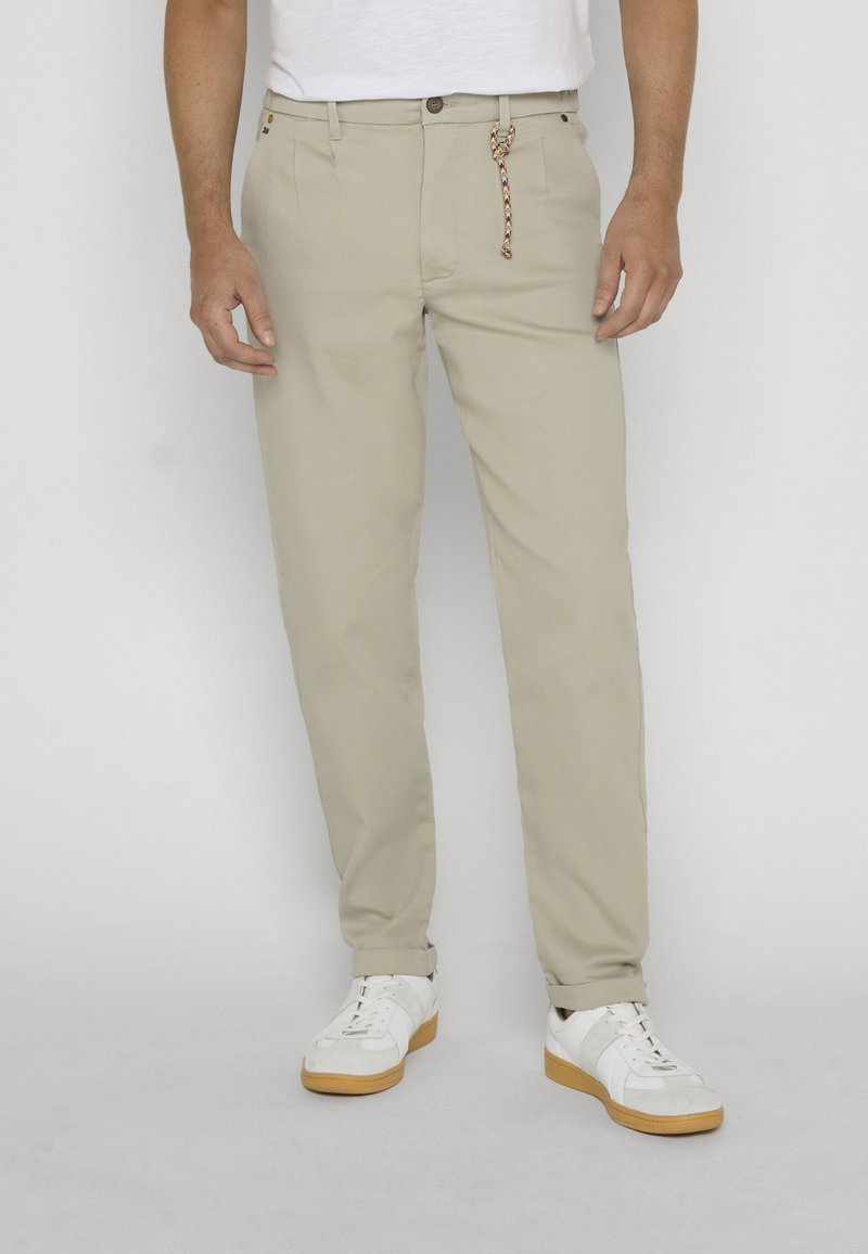Homme portant un pantalon beige coupe droite avec revers roulés, des baskets blanches et une chemise blanche, debout devant un fond neutre.