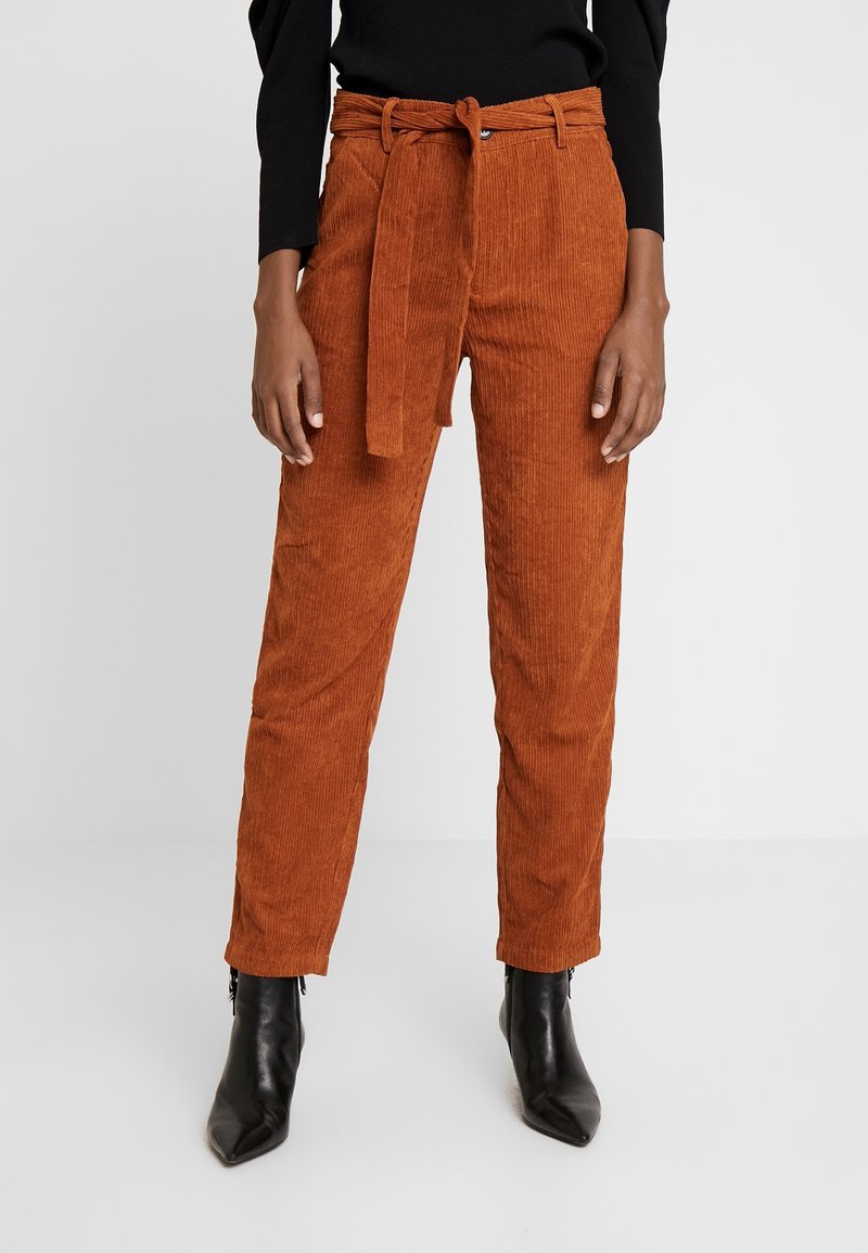 Pantalon en velours côtelé couleur rouille à taille haute avec ceinture nouée à la taille, porté avec des bottines noires à talons et un top noir rentré.