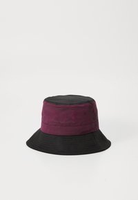 Un chapeau seau avec un dessus bordeaux profond et un bord noir. Tissu texturé avec des détails de couture, comportant des œillets de ventilation sur les côtés.