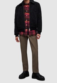 Czarna polarowa kurtka z zamkiem z przodu i ściągaczami, noszona na czerwonej i brązowej koszuli w kratę, beżowych spodniach i czarnych, masywnych butach.