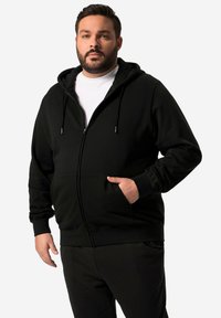 Černá mikina na zip s přední kapsou, kapucí se stahovacími šňůrkami a ribovanými manžetami. Nositá přes bílý tričko s hladkým povrchem.