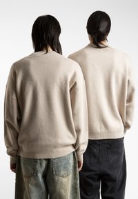 Jerséis de punto beige con un ajuste holgado y puños acanalados, combinados con jeans negros y azules desgastados, mostrados desde la parte de atrás.