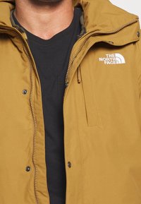 Man som bär en senapsgul The North Face-jacka över en svart V-ringad tröja, jackan delvis öppen som visar detaljer.