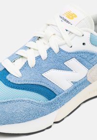 Baskets bleu clair et blanc en daim et mesh, présentant un design en couches, des accents perforés et un logo blanc sur le côté.