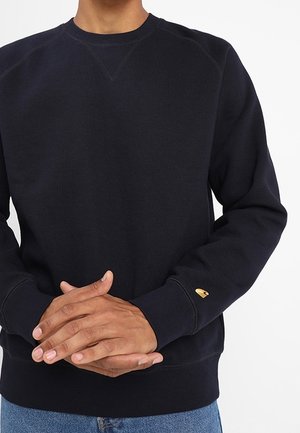Personne portant un sweat-shirt noir uni à manches longues avec des poignets côtelés et un petit logo jaune sur le poignet de la manche gauche.