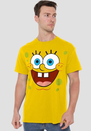 Gelbes Baumwoll-T-Shirt mit einem großen Grafikdruck eines Comic-Charakters, der blaue Augen, einen lächelnden Mund und grüne Akzentpunkte hat.