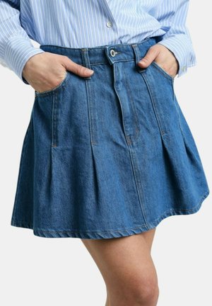Persona che indossa una gonna di jeans blu plissettata e una camicia a righe blu con bottoni, con le mani nelle tasche della gonna.