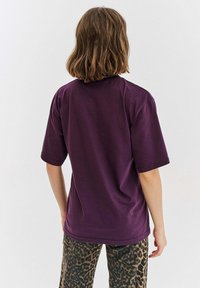 Fialové tričko s krátkým rukávem a kulatým výstřihem, vyrobené z hladké látky. V kombinaci s kalhotami s leopardím potiskem. Zobrazeno zezadu.