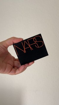 Czarny prostokątny kompakt z pomarańczowym logo "NARS". Gładka matowa powierzchnia z zaokrąglonymi krawędziami. Trzymany w dłoni na jasnym tle.