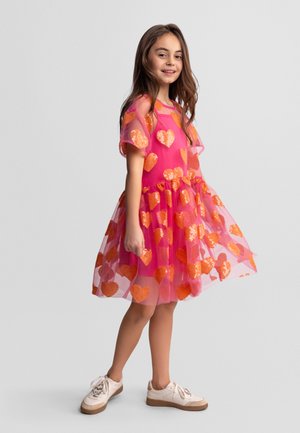 Fille souriante, portant une robe rose vif avec des cœurs en sequins orange et des baskets blanches, debout devant un fond uni.