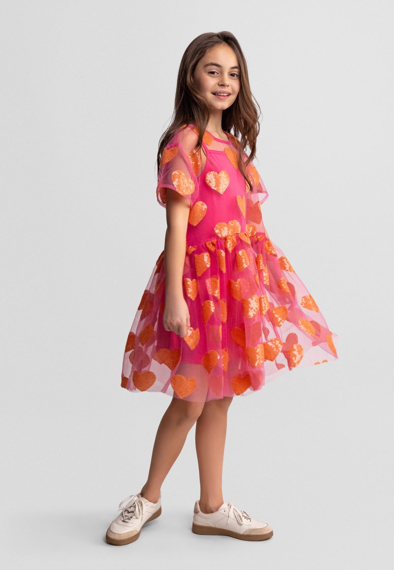 Fille souriante, portant une robe rose vif avec des cœurs en sequins orange et des baskets blanches, debout devant un fond uni.