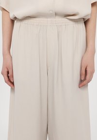 Pantalon beige à taille élastique et jambes larges, porté avec une chemise beige assortie boutonnée, montrant les mains reposant sur les côtés.