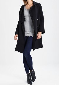 Czarny płaszcz z srebrnymi guzikami, szara wielowarstwowa bluzka, ciemnoniebieskie dżinsy i czarne botki do kostki. Gładka tkanina, dopasowany krój i długie rękawy.