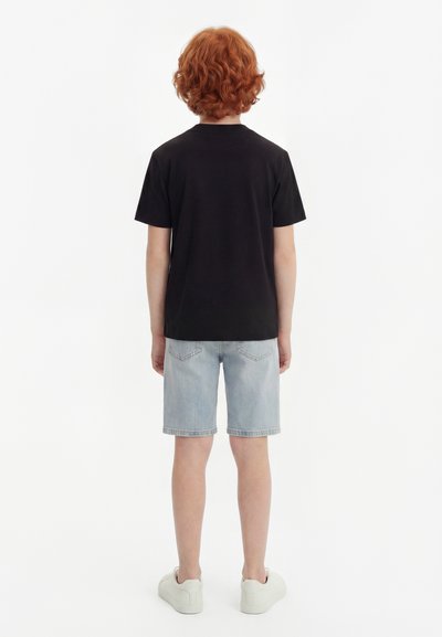 Παιδί με σγουρά κόκκινα μαλλιά που φοράει μαύρο κοντομάνικο μπλουζάκι, ανοιχτό μπλε τζιν σορτς και λευκά αθλητικά παπούτσια, όρθιο με την πλάτη στραμμένη.