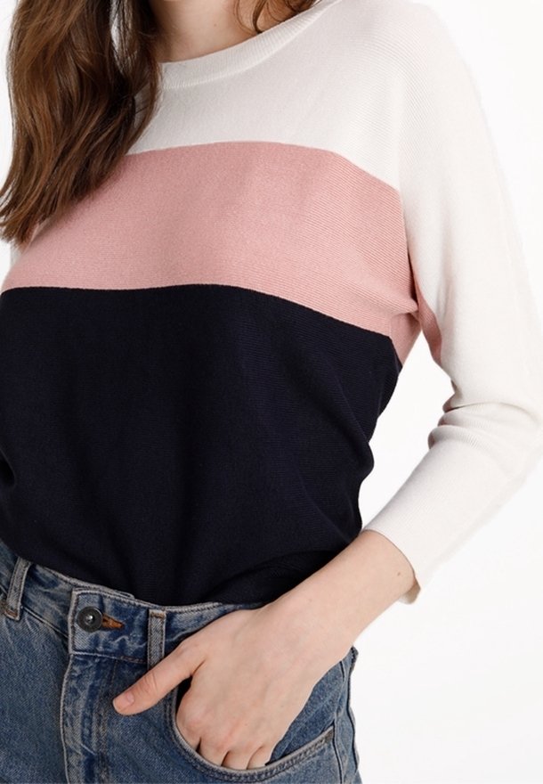 Femme portant un pull color-block blanc, rose et bleu marine rentré dans un jean en denim bleu, la main dans la poche.