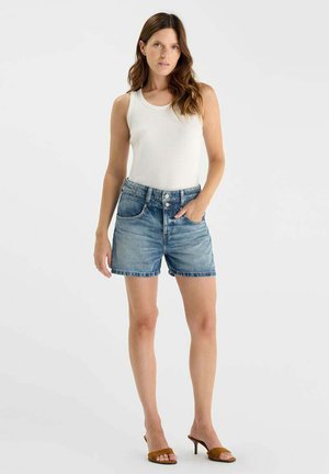 Vrouw staat met hand in zak, draagt een wit mouwloos topje, blauwe denim shorts met knopen en bruine sandalen met hak.