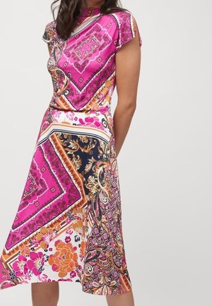 Femme portant une robe midi colorée avec des motifs cachemire et floraux roses, orange et noirs, debout devant un fond clair uni.