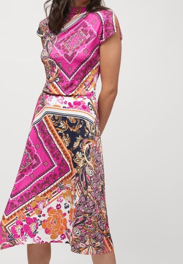 Femme portant une robe midi colorée avec des motifs cachemire et floraux roses, orange et noirs, debout devant un fond clair uni.