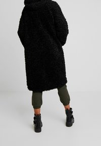 Abrigo largo y esponjoso negro con capucha, usado sobre pantalones cortos verdes oliva y botas de tobillo negras con hebilla. Textura suave y corte relajado.