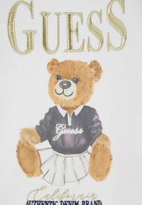 Tričko s medveďom v purpurovom polo tričku s logom "Guess", bielou plisovanou sukňou a zlatými textovými akcentmi na bielom pozadí.