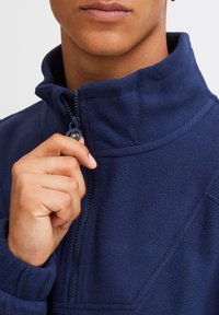 Pullover in fleece blu navy con colletto alto e chiusura a mezzo zip, caratterizzato da una texture morbida e dettagli di cucitura lungo le cuciture.