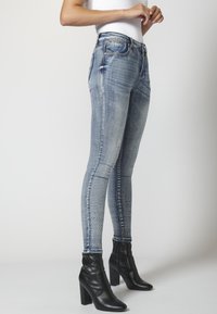 Slim-fit modré džíny s vybledlým povrchem, se střiženými detaily a akcenty z kamínků po stranách, spárované s černými botami na podpatku nad kotníky.