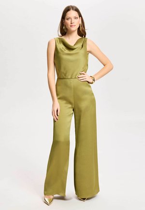 Femme debout portant une combinaison sans manches vert olive à jambes larges avec un col drapé, boucles d’oreilles dorées, bracelet et chaussures dorées pointues.