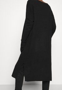 Long black knit cardigan with side slits, soft texture, oversized fit, and ribbed hem; worn over black pants.