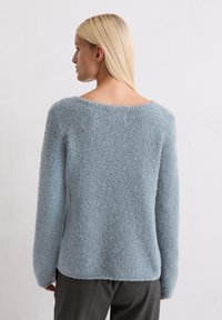 Jasnoniebieski, puszysty sweter z szerokim dekoltem i długimi rękawami, o miękkiej teksturze i lekko luźnym kroju, w zestawie z szarymi spodniami.