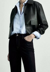 Chaqueta de cuero negra con un cuello con solapa, que presenta un bolsillo en el pecho y grandes botones, combinada con una camisa de rayas azul claro y jeans oscuros.
