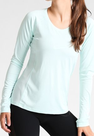 Kobieta ubrana w jasnoniebieską sportową bluzkę z długim rękawem i czarne legginsy, z zawiązanymi włosami, stojąca na jednolitym tle.