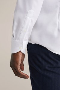 Witte langarmige blouse met een gestructureerde stof, knoopmanchetten en een schuin afgesneden mouwzoom, gecombineerd met donkerblauwe broek.