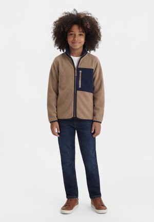 Barn med krøllete hår iført en beige og marineblå fleecejakke med glidelås, mørkeblå jeans og brune joggesko, stående og smilende.