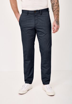 Man wearing dark navy slim-fit trousers, white sneakers, a white shirt, with tattooed right arm visible, standing on a plain white background.