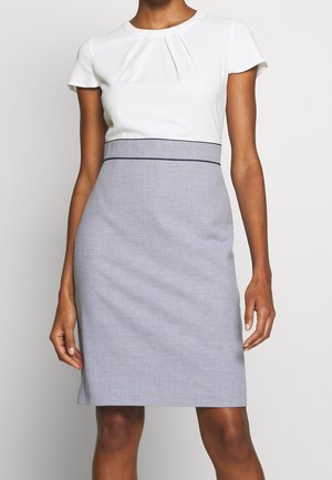 Žena v bílé krátkosleevé šaty s světle šedou sukní a úzkou tmavou linií v pase stojí na bílém pozadí.