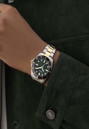 Orologio da polso con quadrante verde, bracciale in acciaio inossidabile argento e oro, lunetta nera e marker luminosi, indossato su una mano con pelle marrone.