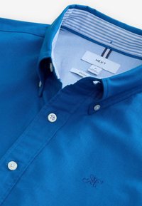 Camisa azul de algodón con botones, que presenta un cuello clásico y botones blancos. Logo bordado sutil en el pecho, cuello interior a rayas.
